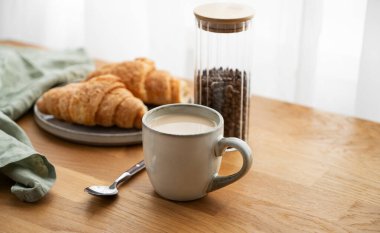 Bir bardak sütlü kahve ve ahşap bir masada taze kruvasan peçete ve kahve çekirdekleri ile pencerenin arka planında. Sabahları sağlıklı kahvaltı kavramı. Boşluğu kopyala.