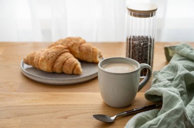 Bir bardak sütlü kahve ve ahşap bir masada taze kruvasan peçete ve kahve çekirdekleri ile pencerenin arka planında. Sabahları sağlıklı kahvaltı kavramı. Boşluğu kopyala.