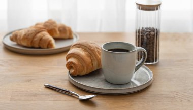 Kahveli espresso ve kruvasan tabakta, ahşap bir masada, pencerenin arka planına karşı. Sabahları sakin bir kahvaltı kavramı var. Ön görünüm ve boşluk kopyala.
