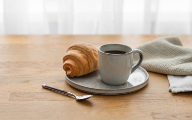 Kahveli espresso ve kruvasan bir tabakta, tahta bir masanın üzerinde ve peçeteyle pencerenin arka planında. Sabahları sakin bir kahvaltı kavramı var. Ön görünüm ve boşluk kopyala.