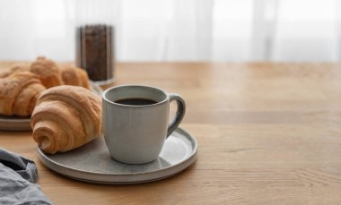 Bir bardak taze demlenmiş kahve ve kruvasan ahşap bir masanın üzerindeki bir tabakta pencerenin arka planına karşı. Sabahları sakin bir kahvaltı kavramı var. Ön görünüm ve boşluk kopyala.
