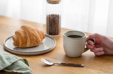 Elinde bir bardak taze demlenmiş kahveyle ahşap bir masanın üzerinde bir pencerenin arka planında kruvasan vardı. Sabahları sakin bir kahvaltı kavramı..