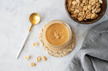 Gri arka planda kremalı fıstık ezmeli cam kavanoz. Kavrulmuş fındık, kaşık ve peçete. Doğal ev yapımı fıstık ezmesi konsepti. Üst görünüm ve kopyalama alanı.