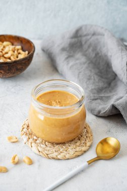 Gri arka planda kremalı fıstık ezmeli cam kavanoz. Kavrulmuş fındık, kaşık ve peçete. Doğal ev yapımı fıstık ezmesi konsepti. Ön görünüm ve boşluk kopyala.