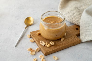 Ahşap bir zeminde üzerinde kremalı fıstık ezmesi olan cam kavanoz. Kavrulmuş fındık, kaşık ve peçeteyle birlikte. Doğal ev yapımı fıstık ezmesi konsepti. Boşluğu kopyala.