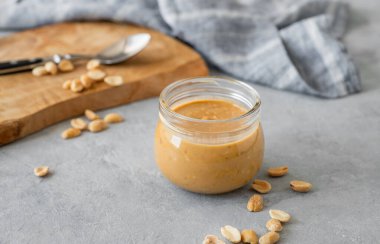 Gri arka planda kremalı fıstık ezmesi, saçılmış fındık, ahşap tahta ve peçeteyle dolu cam kavanoz. Doğal ev yapımı fıstık ezmesi konsepti. Boşluğu kopyala.