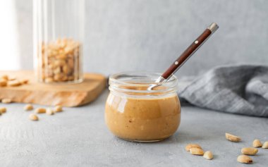 Gri arka planda kremalı fıstık ezmeli cam kavanoz dağınık kavrulmuş fındık, ahşap tahta ve güneş ışığı. Doğal ev yapımı fıstık ezmesi konsepti. Ön görünüm ve boşluk kopyala.