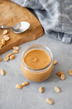 Gri arka planda kremalı fıstık ezmesi, saçılmış fındık, ahşap tahta ve peçeteyle dolu cam kavanoz. Doğal ev yapımı fıstık ezmesi konsepti. Üst görünüm.