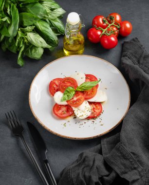 Dilimlenmiş domatesli Caprese salatası, mozzarella ve koyu arkaplanda zeytinyağı ve bitkilerle dolu bir tabakta fesleğen. Sağlıklı Akdeniz mutfağı kavramı. 