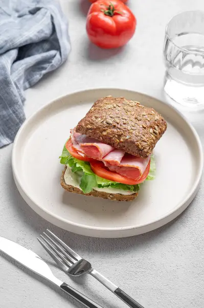 Prosciutto, peynir, domates, marul ve sostan yapılmış tahıllı sandviç hafif bir arka planda bir tabakta, bir bardak su, peçete ve çatalla. Sağlıklı ve diyet kahvaltı konsepti. Üst görünüm.