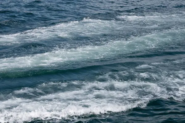 Çalkalanan okyanus dalgaları, gün ışığında hareket ve deniz enerjisini temsil eden derin mavi deniz suyunun yüzeyinde köpüklü bir iz bırakır..