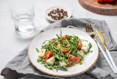 Kiraz domatesli sağlıklı roka salatası, mozzarella ve hafif dokulu arka planda bir tabakta pesto sosu, bir bardak suyla. Taze ve sağlıklı Akdeniz gıda kavramı.