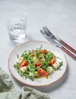 Kiraz domatesli sağlıklı roka salatası, mozzarella ve hafif dokulu arka planda bir tabakta pesto sosu, bir bardak suyla. Taze ve sağlıklı Akdeniz gıda konsepti. Boşluğu kopyala. 