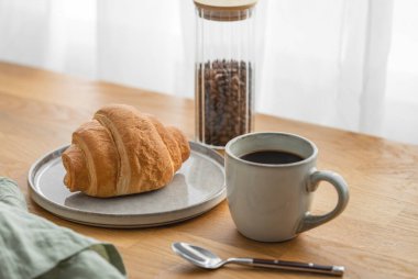 Bir bardak taze demlenmiş kahve, ahşap bir masanın üzerinde ve arka planda kruvasanlar var. Sabahları sakin bir kahvaltı kavramı..