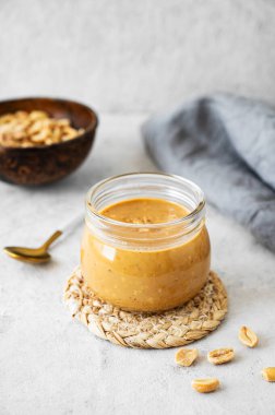 Ahşap bir zeminde üzerinde kremalı fıstık ezmesi olan cam kavanoz. Kavrulmuş fındık, kaşık ve peçeteyle birlikte. Doğal ev yapımı fıstık ezmesi konsepti. Boşluğu kopyala.