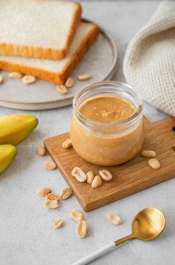 Gri arka planda kremalı fıstık ezmesi, muz ve taze ekmekle dolu cam kavanoz. Doğal ev yapımı fıstık ezmesi konsepti.