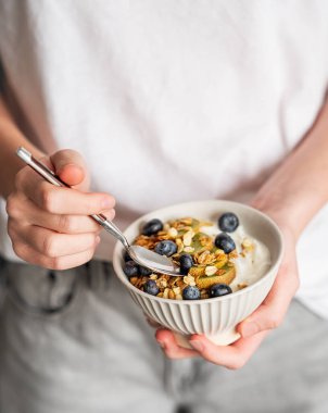 Bir kase granola, Yunan yoğurdu, kivi, yabanmersini ve kaşığı beyaz tişörtlü kadının ellerinde. Sütlü kahvaltı konsepti, sabah ışığında yulaf ezmesi..
