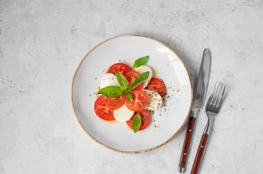 Dilimlenmiş domatesli Caprese salatası, mozzarella ve doğal ışık altında çatal ve bıçakla beyaz arka planda bir tabakta fesleğen. Sağlıklı Akdeniz mutfağı konsepti. Üst görünüm ve kopyalama alanı.