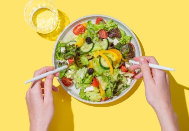 Elleri sebze salatasıyla marul, zeytin ve beyaz peynirle karıştırılmış sarı arka planda çatal ve kaşık ile bir bardak su. Pankart reklamı veya menü düzeni için taze ve doğal sağlıklı gıda kavramı.