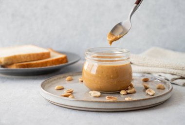 Cam kavanozdaki kaşıktan akan fıstık ezmesi hafif arka planda tuzlu fındık ve ekmek kızartmalarıyla birlikte. Kahvaltıda tatlı sandviçler için lezzetli ve sağlıklı atıştırmalık kavramı. Ön görünüm ve boşluk kopyala.