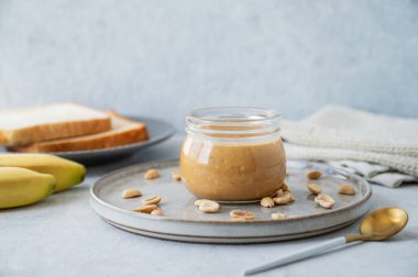 Hafif arka planda cam kavanozda tuzlanmış fındık, muz ve ekmek kızartmasıyla fıstık ezmesi. Kahvaltıda tatlı sandviçler için lezzetli ve sağlıklı atıştırmalık kavramı. Ön görünüm ve boşluk kopyala.