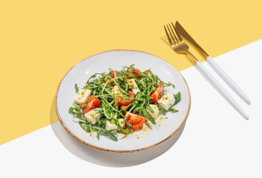 Kiraz domatesli taze roka salatası, mozzarella sosu beyaz sarı arka planda, çatal bıçaklı pesto sosu. Taze ve sağlıklı Akdeniz mutfağı kavramı. Boşluğu kopyala.