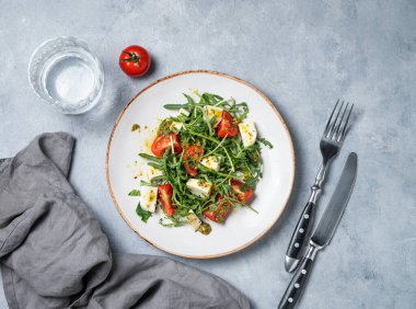 Kiraz domatesli Arugula salatası, mozzarella ve bir bardak su ve çatal bıçakla arka planda pesto sosu. Taze ve sağlıklı Akdeniz gıda konsepti. Üst görünüm, alanı kopyala.
