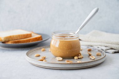 Cam kavanozda fıstık ezmesi, hafif arka planda kaşık, tuzlu fındık ve ekmek kızartması. Kahvaltıda tatlı sandviçler için lezzetli ve sağlıklı atıştırmalık kavramı. Ön görünüm ve boşluk kopyala.
