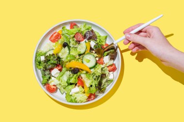 El ele tutuşan, salatalı, marullu, zeytinli ve sarı arka planda peynirli, çatallı sebze salatası. Afiş reklamı ya da menü düzeni için taze ve doğal sağlıklı gıda konsepti. Üst görünüm.