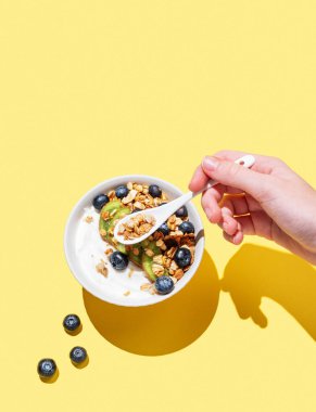 Sarı arka planda mısır gevreği, kivi ve yaban mersinli kaşık dolusu yoğurt. Sağlıklı ve besleyici kahvaltı konsepti, kafe menüsü reklam afişi. Üst görünüm, alanı kopyala