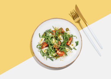 Kiraz domatesli taze roka salatası, mozzarella sosu beyaz sarı arka planda, çatal bıçaklı pesto sosu. Taze ve sağlıklı Akdeniz mutfağı kavramı. Üst görünüm ve kopyalama alanı.