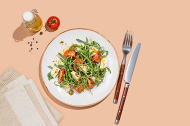 Çeri domatesli taze roka salatası, mozzarella, bej arka planda peçete ve çatal bıçaklı pesto sosu. Taze ve sağlıklı Akdeniz mutfağı kavramı. Üst görünüm ve kopyalama alanı.