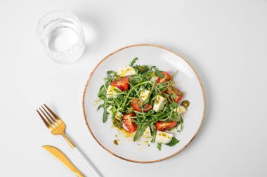Çeri domatesli taze roka salatası, mozzarella, beyaz zemin üzerinde pesto sosu, bir bardak su ve çatal bıçak takımı. Taze ve sağlıklı Akdeniz mutfağı kavramı. Üst görünüm ve kopyalama alanı.