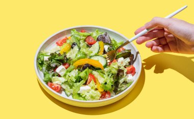 El ele tutuşan, salatalı, marullu, zeytinli ve sarı arka planda peynirli, çatallı sebze salatası. Afiş reklamcılığı veya menü düzeni için taze ve doğal sağlıklı gıda konsepti.