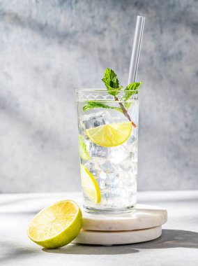Uzun bardakta mojito kokteyli, kireç, nane ve buzla mermer tezgahın üzerinde gölgeli, hafif bir arka planda. Turunçgil meyveleriyle ferahlatıcı limonata içeceği konsepti. Ön görünüm, boşluğu kopyala