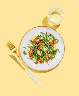 Çeri domatesli taze roka salatası, mozzarella, sarı zemin üzerinde pesto sosu, bir bardak su ve çatal bıçak takımı. Taze ve sağlıklı Akdeniz mutfağı kavramı. Üst görünüm ve kopyalama alanı.