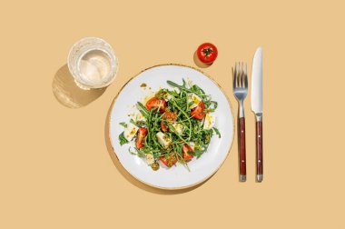 Çeri domatesli taze roka salatası, mozzarella, sarı zemin üzerinde pesto sosu, bir bardak su ve çatal bıçak takımı. Taze ve sağlıklı Akdeniz mutfağı kavramı. Üst görünüm ve kopyalama alanı.