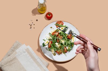 El ele tutuşan taze roka salatası, kiraz domatesleri, mozzarella, bej arka planda pesto sosu. Taze ve sağlıklı Akdeniz mutfağı kavramı. Üst görünüm ve kopyalama alanı.