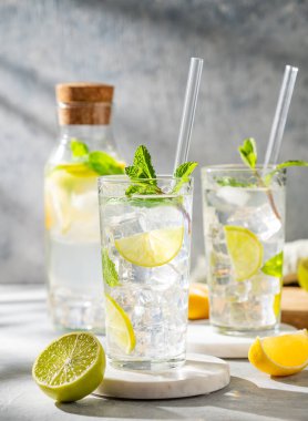 İki bardak mojito kokteyli. Taze limonlu, naneli ve buzlu. Arka planda hafif bir şişe ve gölge var. Turunçgil meyveli yaz ferahlatıcı içecek konsepti. Ön görünüm.