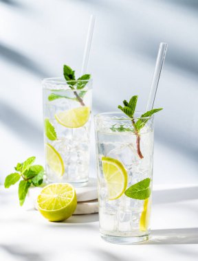 İki bardak mojito kokteyli, limon, nane, buz ve gölgeli beyaz mavi arka planda tüp. Turunçgil meyveleriyle ferahlatıcı limonata içeceği konsepti. Ön görünüm, boşluğu kopyala