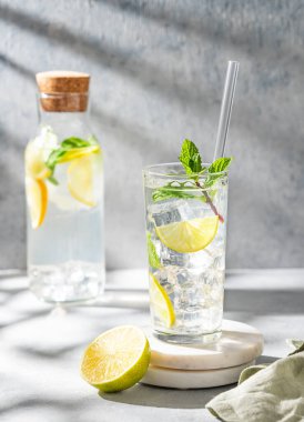 Limonlu, naneli ve buzlu bir bardakta mojito kokteyli mermer tezgahın üzerinde, arka planda hafif bir şişe ve gölgeyle. Turunçgil meyveleriyle ferahlatıcı limonata içeceği konsepti. Ön görünüm.