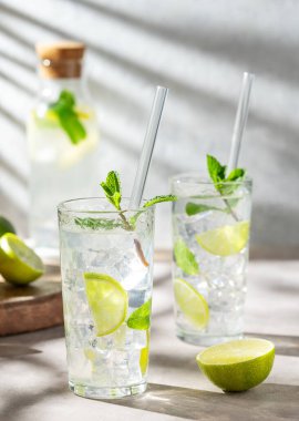 İki bardak mojito kokteyli. Taze limonlu, naneli ve buzlu. Arka planda hafif bir şişe ve gölge var. Turunçgil meyveli yaz ferahlatıcı içecek konsepti. Ön görünüm.