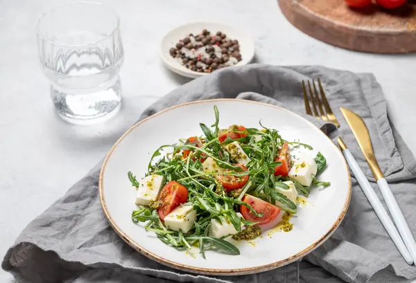 Kiraz domatesli sağlıklı roka salatası, mozzarella ve hafif dokulu arka planda bir tabakta pesto sosu, bir bardak suyla. Taze ve sağlıklı Akdeniz gıda kavramı.