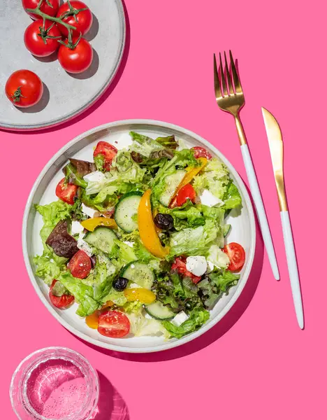 Sebze salatası, marul, zeytin ve pembe arka planda beyaz peynir ve bir bardak su ve çatal bıçak. Afiş reklamı ya da menü düzeni için taze ve doğal sağlıklı gıda konsepti. Üst görünüm.