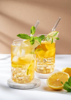 İki bardak buzlu çay, limon, nane ve buz beyaz bej arka planda, güneş ışığıyla mermer bir stantta. Bir yaz günü kokteyl ya da limonata konsepti. Ön görünüm, boşluğu kopyala.