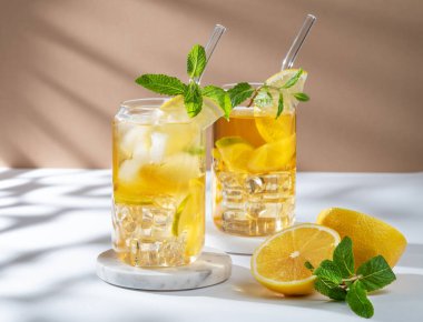 İki bardak buzlu çay, limon, nane ve buz beyaz bej arka planda, güneş ışığıyla mermer bir stantta. Bir yaz günü kokteyl ya da limonata konsepti. Ön görünüm, boşluğu kopyala.