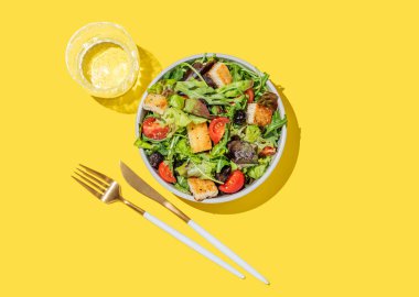 Sebzeli salata, marul, domates, kızarmış ekmek ve zeytinler sarı arka planda bir kasede, bir bardak su ve çatal bıçakla. Reklam menüsü için sağlıklı diyet yemekleri konsepti. Üst görünüm, alanı kopyala.