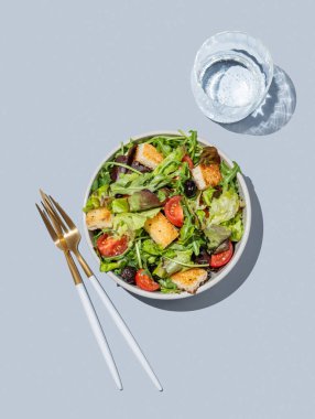 Marul, domates, kızarmış ekmek ve zeytinli sebze salatası mavi arka planda bir kasede, bir bardak su ve çatal bıçakla. Reklam menüsü için sağlıklı diyet yemekleri konsepti. Üst görünüm, alanı kopyala.