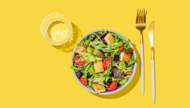 Sebzeli salata, marul, domates, kızarmış ekmek ve zeytinler sarı arka planda bir kasede, bir bardak su ve çatal bıçakla. Reklam menüsü veya afiş için sağlıklı diyet gıda konsepti. Üst görünüm