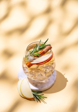 Elma kokteyli ya da buzlu bir bardakta elma şarabı, tarçın ve biberiye mermer bir tezgahta gölgeli sarı bir arka planda. Reklamcılık ya da bar menüsü afişi için canlandırıcı içecek konsepti. Boşluğu kopyala